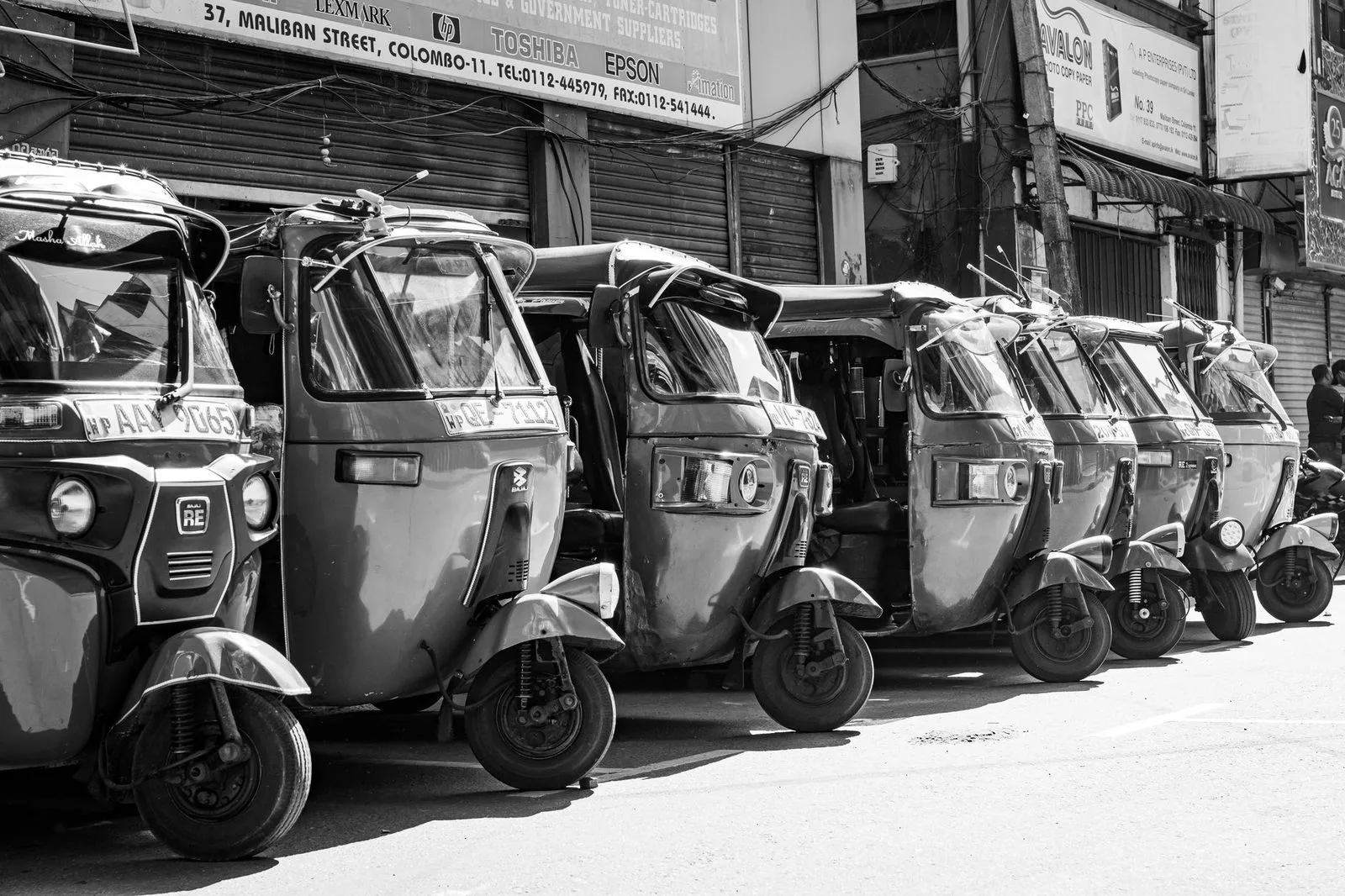Stock photograph of several auto-rickshaws parked in a line beside a road; the vehicles and people are unaffiliated with Wanhoo